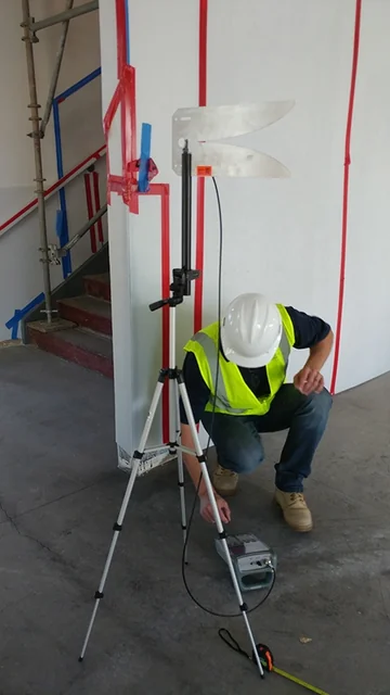 A technician in a hard hat and high-visibility vest crouches on bare concrete to read a handheld meter, while a tripod-mounted twin-blade antenna beside him is aimed at a tall white wall panel; scaffolding and taped layout lines stand behind, in a building shell still under construction.